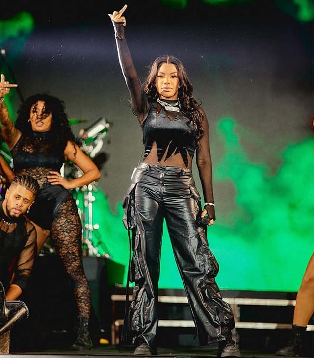 Ludmilla, de calça de couro preta e top, aponta para o alto no palco, com bailarinas atrás e fumaça verde
