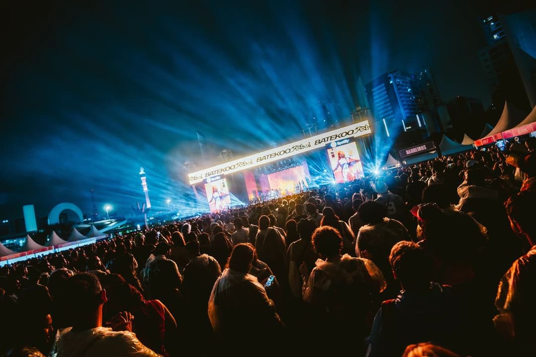 Multidão à noite diante de um palco iluminado por fachos azuis, com o letreiro BATEKOO Festival, dois telões e prédios ao fundo