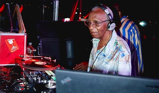 Senhor de cabelo branco, fones de ouvido e camisa estampada mexe nos toca-discos de uma cabine de DJ