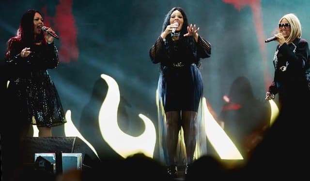 Três cantoras de roupa preta no palco, diante de um telão com chamas desenhadas