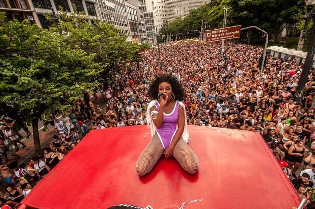 Uma dançarina de maiô lilás, ajoelhada no teto de um trio, olha para uma avenida tomada pelo público