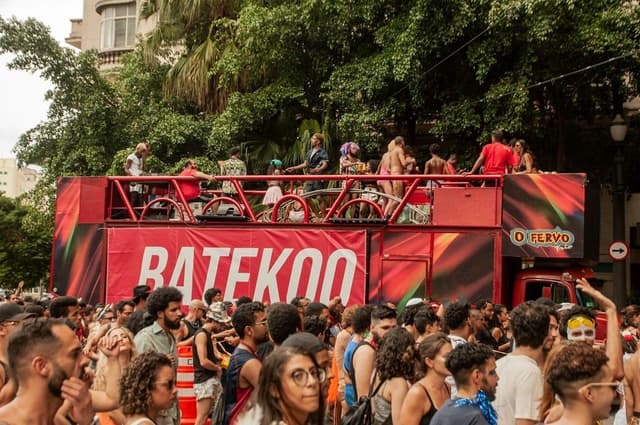 Trio elétrico vermelho com o nome BATEKOO cercado por uma multidão numa rua arborizada do centro de São Paulo
