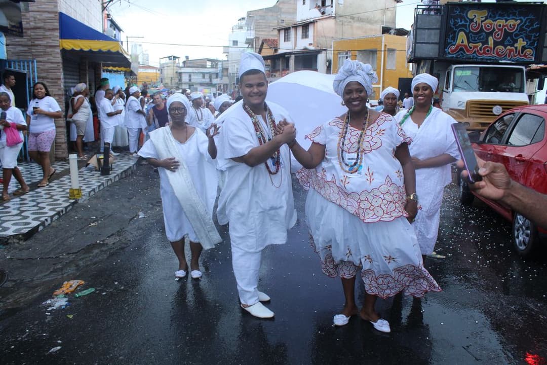 Mulheres e homens vestidos de branco, com turbantes e saias rodadas, caminham por uma rua de bairro em Salvador