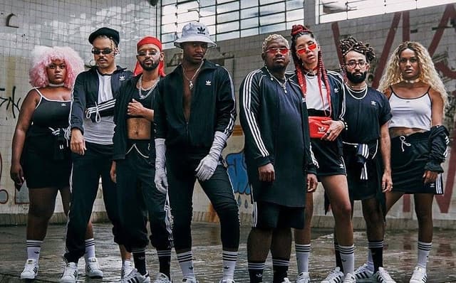 Nove jovens negros com roupas esportivas pretas e brancas posam lado a lado num galpão de paredes grafitadas