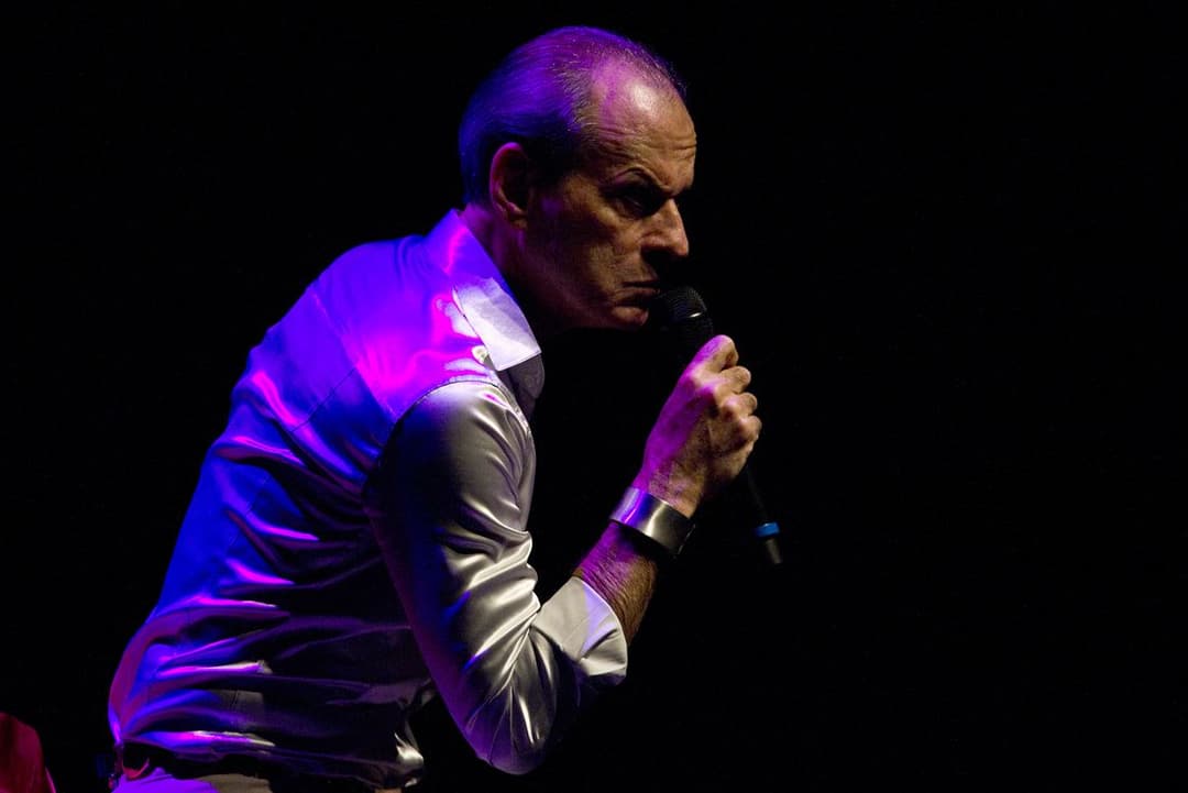 Ney Matogrosso, de camisa brilhante, canta curvado ao microfone sob luz roxa