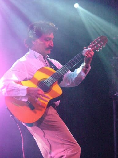 Belchior, de bigode e camisa branca, toca violão num palco sob luz colorida