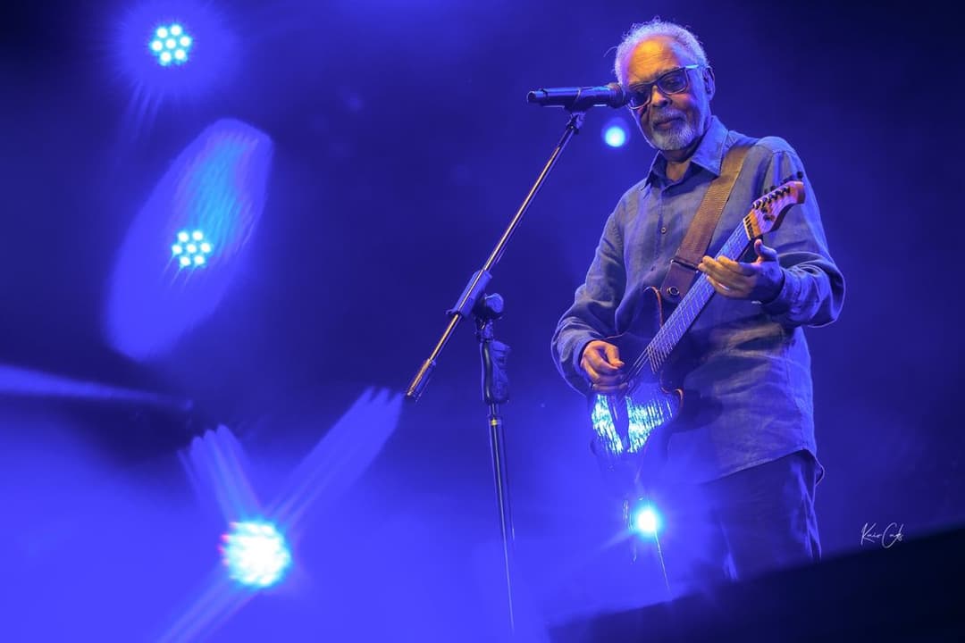 Gilberto Gil, de óculos e camisa azul, toca guitarra diante do microfone sob luzes azuis
