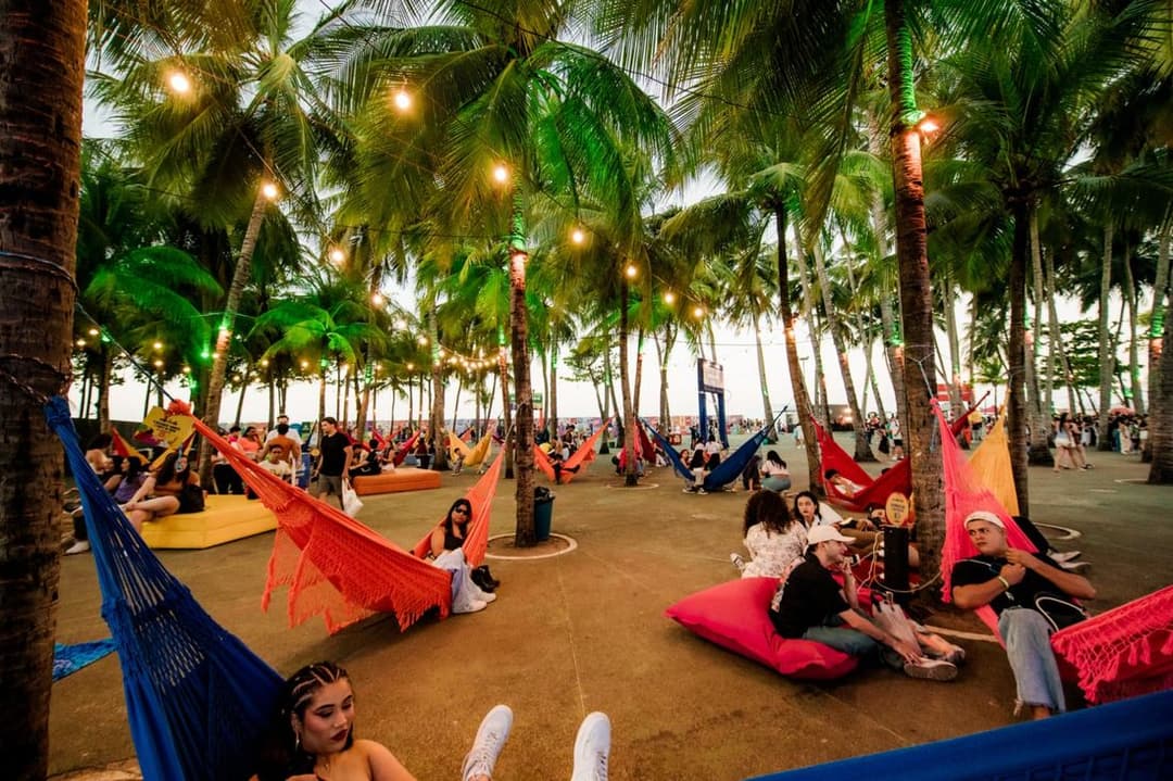 Redes coloridas e almofadas sob coqueiros iluminados por cordões de lâmpadas, com pessoas descansando