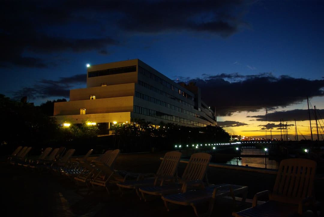 Prédio de hotel iluminado ao anoitecer, com espreguiçadeiras vazias à beira da piscina em primeiro plano