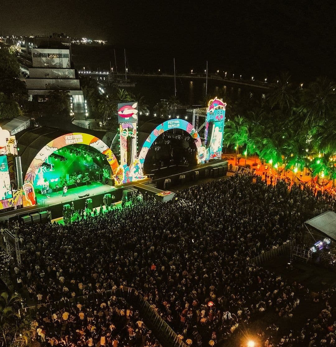 Vista aérea noturna do festival: dois palcos em arco, iluminados, diante de uma multidão, com coqueiros, prédios e o mar ao fundo