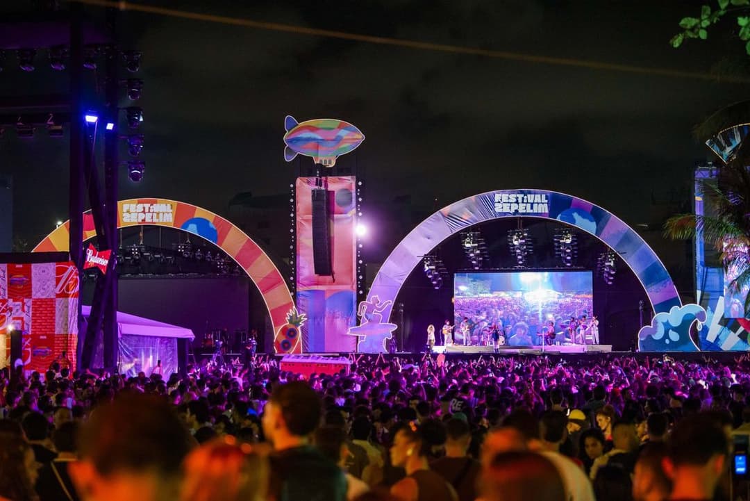Multidão à noite diante de um palco com arcos coloridos e telão, com um zepelim decorativo no alto da estrutura